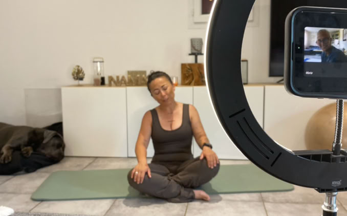 Professeure de Doga assise sur son tapis devant un téléphone pour guider une séance à distance.