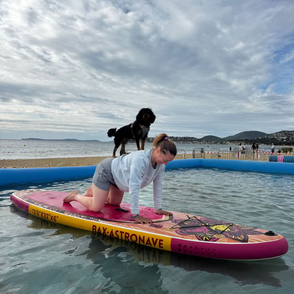 Femme pratiquant le yoga sur une planche de paddle avec un chien debout derrière elle.